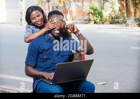 young man sitting outdoors working with laptop while his girlfriend covers his eyes with his hands smiling Stock Photo