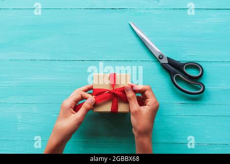 Christmas background with gift box and scissors on teal blue wooden table. Preparation for holidays. Top view, flat lay. Girl's hands holding gift box Stock Photo