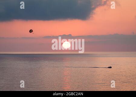 Parasailing behind a boat at sunset over the sea with the sun goes down. Silhouette of a person with parachute against evening sky with clouds. Beautiful seascape. Stock Photo