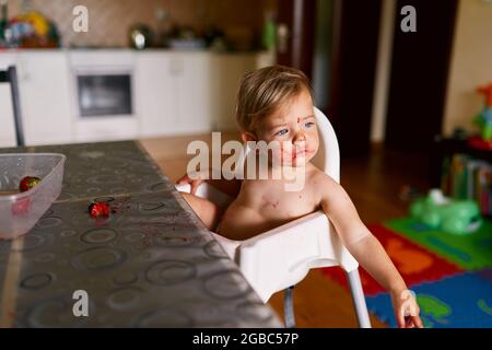 Kid in a diaper with a dirty face sits on a high chair Stock Photo