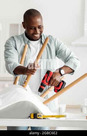 Manual job at home. Happy millennial black man assembling furniture with electric screwdriver in kitchen interior, crop Stock Photo