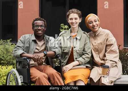 Group of positive young multi-ethnic friends sitting on bench and drinking coffee with handicapped man outdoors Stock Photo