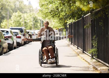 Happy excited young African-American disabled man in wheelchair pushed by running woman outstretching arms and feeling freedom Stock Photo