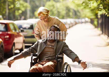 Positive young African-American disabled man chatting with friend who pushing him in wheelchair along street Stock Photo