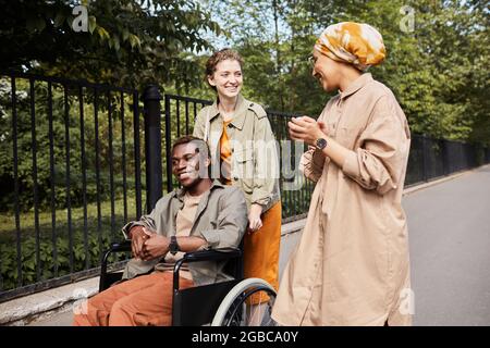 Young multi-ethnic friends talking during stroll: Caucasian woman pushing wheelchair with disabled man while talking to Arabian friend Stock Photo