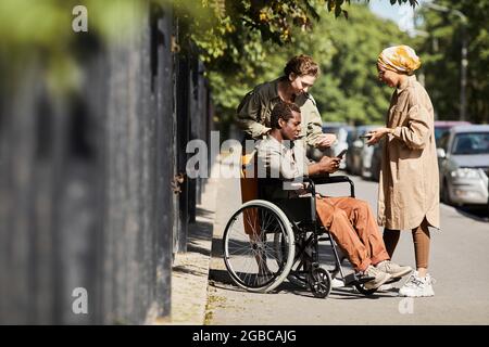 Content disabled young African-American man in wheelchair showing video on social media to female friends on street Stock Photo