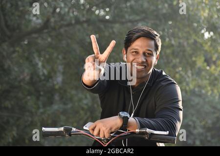 young happy Indian man sitting on cycle showing Victory sign and listen to music Stock Photo