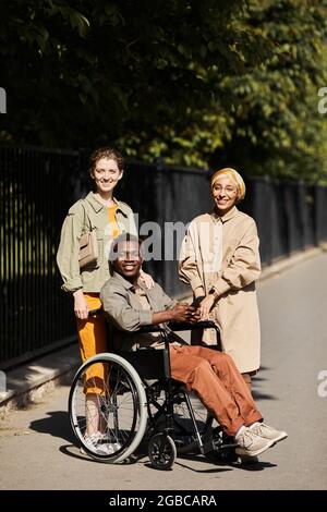 Portrait of smiling young women standing near handsome handicapped man in wheelchair outdoors Stock Photo