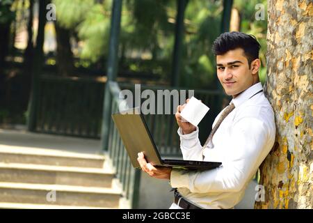 young Indian man drinking coffee and working on laptop at park, young Indian business man Stock Photo