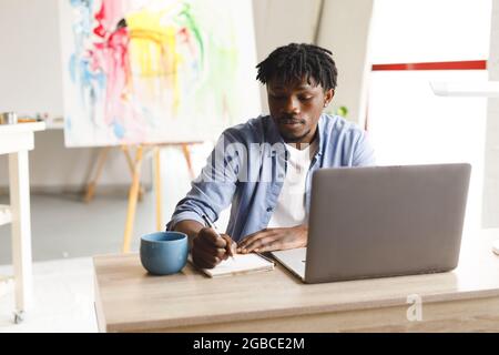 African american male painter at work using laptop and taking notes in art studio Stock Photo