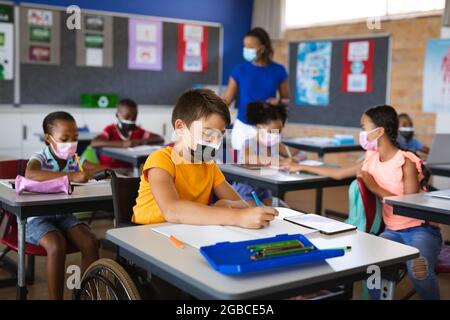 Disabled caucasian boy wearing face mask studying while sitting on wheelchair at elementary school