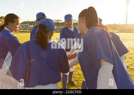 Diverse group of female baseball players warming up on field at sunrise, stacking hands Stock Photo