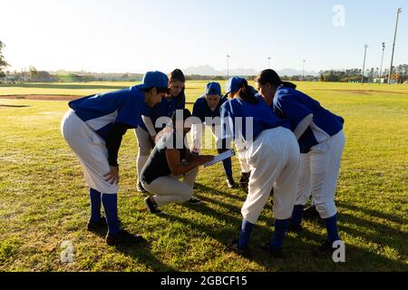 Diverse group of female baseball players standing in huddle around squatting coach on field Stock Photo