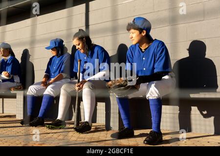 Diverse group of female baseball players sitting on bench in sun, waiting to play Stock Photo
