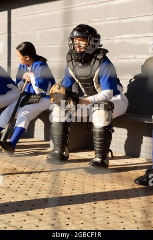 Diverse group of female baseball players sitting on bench, waiting to play game Stock Photo