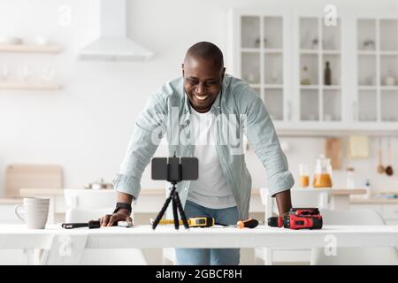 Recording vlog about repair and construction tools. Happy black man, talking to cellphone webcam in kitchen interior Stock Photo