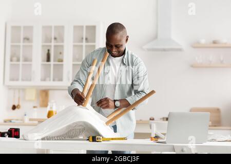 Online instructions of furniture assembly. Happy black handyman screwing detail to chair, using laptop computer Stock Photo