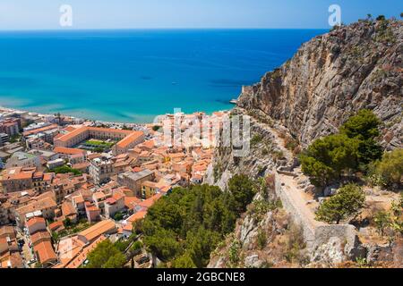 Cefalù, Palermo, Sicily, Italy. City rooftops dwarfed by the towering cliffs of La Rocca, the turquoise waters of the Tyrrhenian Sea beyond. Stock Photo