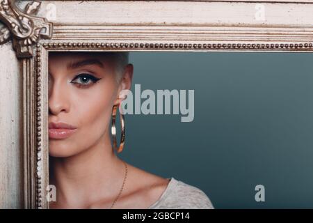 Millenial young woman with short blonde hair on gilded picture frame. Cropped face of beautiful mysterious girl in gold baroque frame corner. Isolated on blue background in studio. Copy space. Stock Photo