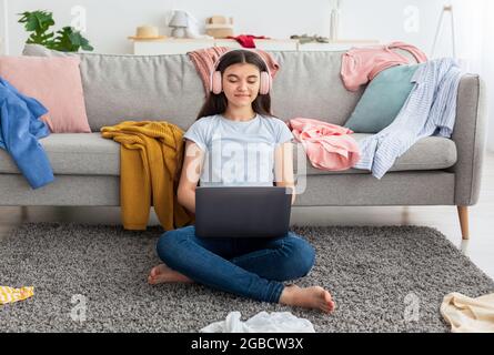 Web based schooling. Lovely Indian teen girl in headphones speaking to friends on webcam, using laptop in messy room Stock Photo