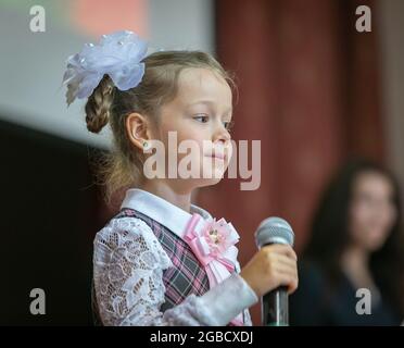 On September 1, a girl in the school hall reads a verse into a microphone. Girl with pigtails and bows. Moscow, Russia, September 2, 2019 Stock Photo