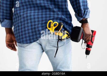 Tool for drilling and fixing. African american man holding electric drill, measuring tape and scissors in pocket Stock Photo