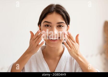 Cheerful Woman Applying Facial Cream Moisturizing Skin Posing In Bathroom Stock Photo