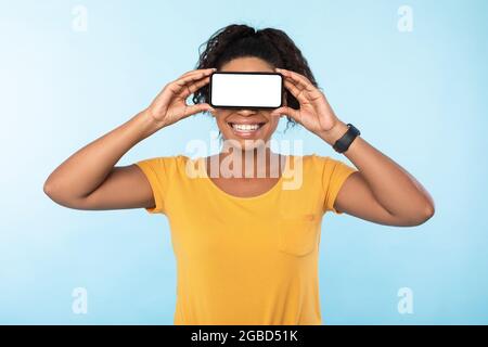 Happy black woman covering eyes with blank smartphone, recommending mobile app, advertising new website, mockup Stock Photo