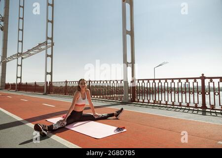 Attractive positive woman does stretching exercise sitting on contemporary bridge Stock Photo