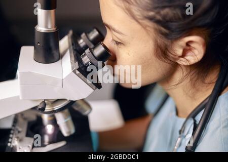 Asian lady doctor looks into contemporary microscope researching sample on slide in hospital Stock Photo