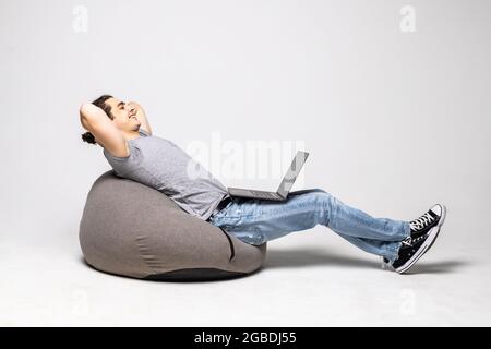 Young man with laptop sitting on beanbag chair against white background Stock Photo