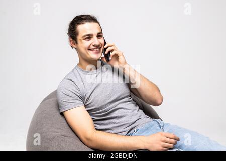 Handsome young man wearing a casual outfit, sitting on a bean bag, holding his phone talking to someone, looking away, isolated on white background Stock Photo