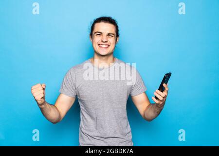Emotional excited screaming man isolated over blue wall background play games by mobile phone make winner gesture. Stock Photo