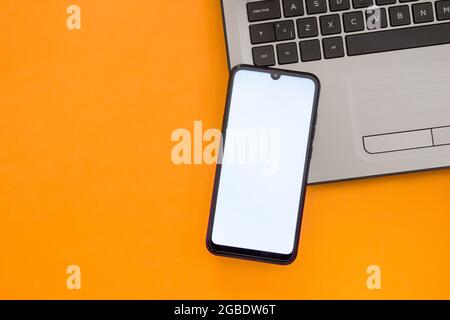 Smartphone with blank white screen on the notebook keyboard. Mockup phone. Top view. Flat lay. Isolated on orange background. Stock Photo