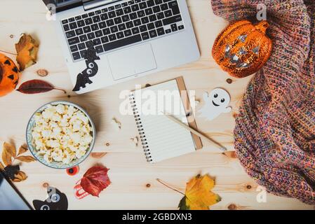 Top view Blank notebook on wooden table with laptop, popcorn, Halloween decor, lighting pumpkin, autumn leaves and warm plaid. Planning list of films Stock Photo