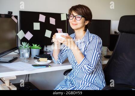 Woman in glasses drinking coffee on the break to relax during work on her home office workplace. Satisfied with work done. Taking time for energy Stock Photo