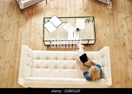 Top view of mature woman in casual wear using tablet pc while working from home, doing some paperwork, sitting on a couch in modern apartment Stock Photo