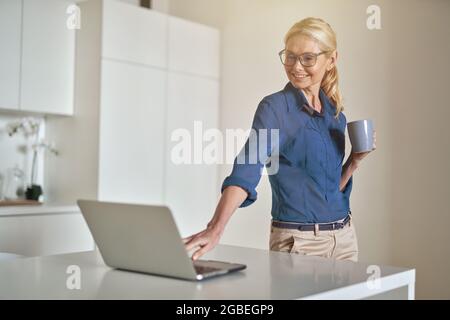 Smiling blonde woman in glasses having a cup of coffee and using laptop in the morning, while working from home Stock Photo