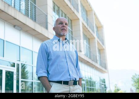 Troubled senior man staring up in angst at the sky as he stands with hands in pockets in front of an office block in a low angle close up view Stock Photo