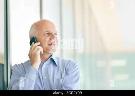 Senior businessman chatting on his smartphone in a high key portrait as he stands leaning against glass windows of a commercial building or office blo Stock Photo