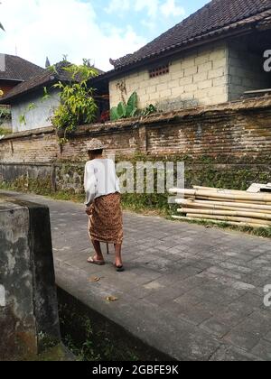 An elderly man wearing a palm frond hat and colorful sarong walks past a pile of bamboo poles in a narrow paved lane in Bali, Indonesia. July 23, 2021 Stock Photo