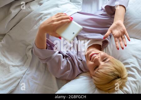 Young woman in bed during online video call. Online meeting with friends. She waving hand and saying hello after awakening. Stock Photo