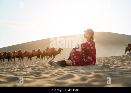 asian woman in red dress sitting in desert looking at view with caravan of camels and huge sand dune in background Stock Photo