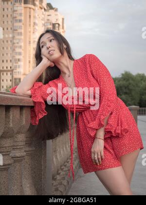 Attractive smiling Asian girl in red dress posing in the city near the balustrade. Stock Photo