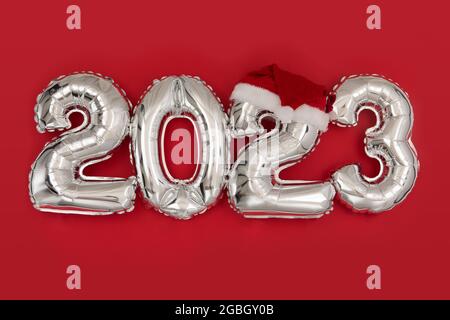 2023 inflatable balloons on red background with santa hat. Happy New year 2023 celebration. Silver foil balloons numeral 2023. Postcards and posters C Stock Photo