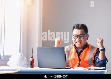 Smiling engineer sitting at his desk celebrating with his hands in the air Stock Photo