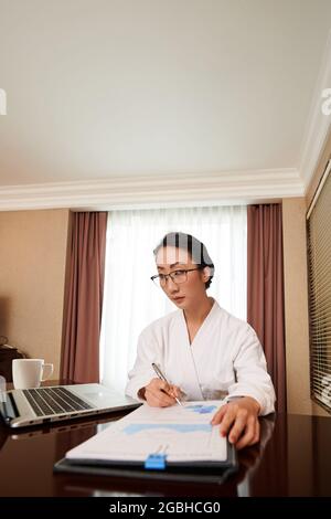 Concentrated young Asian businesswoman in eyeglasses editing data and looking at laptop screen while working during vacation Stock Photo