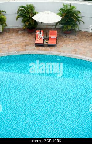 Above view of young Asian woman making video call and shooting video for social media while resting near pool Stock Photo
