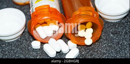 Horizontal view of pills spilling out of two prescription drug bottles on a black counter with the white tops on the side. Stock Photo