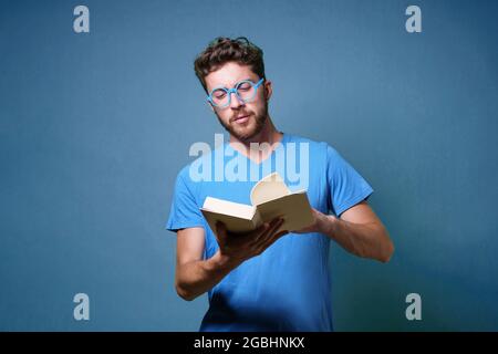 Portrait of a young student man with eyeglasses reading a book. Portrait isolated on blue colored background. Studio shot. Stock Photo